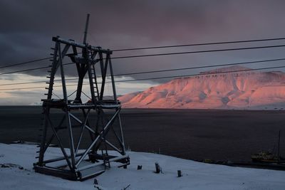 Electricity pylon by snowcapped mountains against sky at sunset