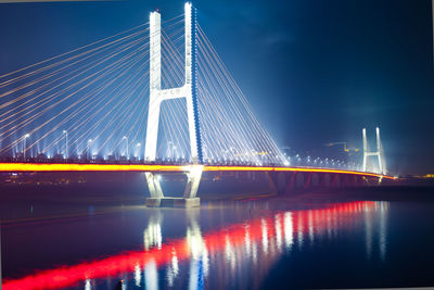 Illuminated suspension bridge over river against sky at night