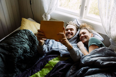 High angle view of smiling friends watching movie over digital tablet while lying on bed in cottage