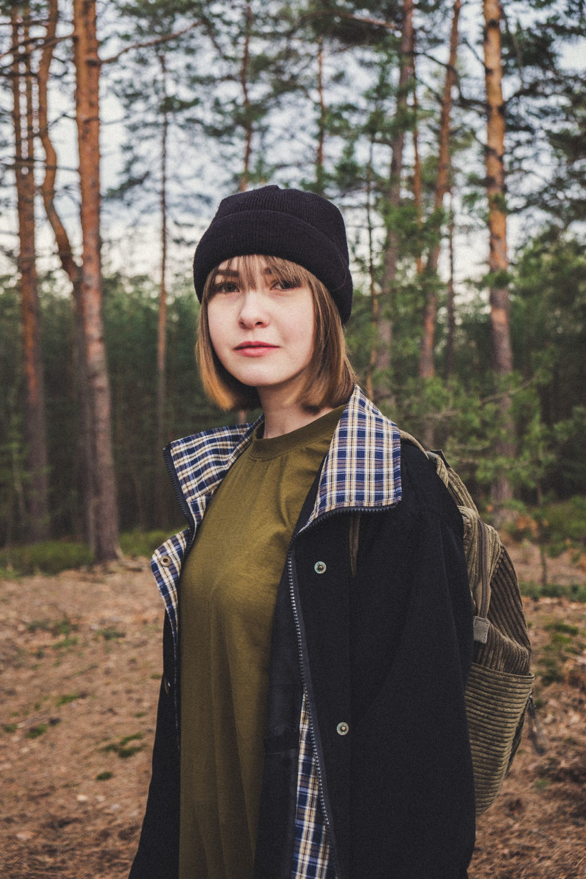 tree, forest, land, clothing, plant, one person, young adult, front view, standing, portrait, real people, leisure activity, warm clothing, waist up, knit hat, young women, winter, day, nature, hairstyle, woodland, outdoors, teenager