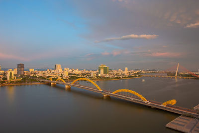 Illuminated bridge over river by buildings against sky in city