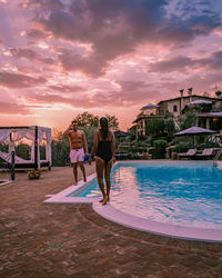 Full length of couple standing by swimming pool against sky during sunset