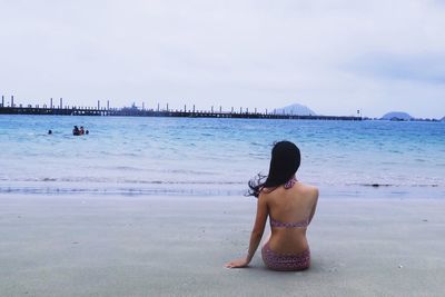 Rear view of woman sitting on shore at beach against sky