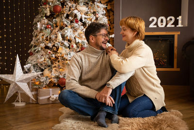 Smiling mature couple sitting by christmas tree at home