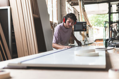 Male carpenter measuring plank using tape measure on workbench in workshop