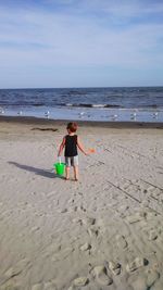 Rear view of boy holding shovel and pail while standing at beach against sky