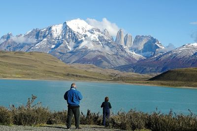 Man and boy standing at lakeshore against snowcapped mountains
