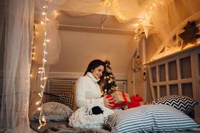 Side view of young woman holding christmas present while sitting on bed at home