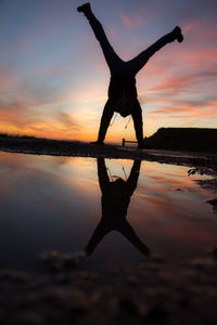 Silhouette man doing handstand by lake against sky during sunset