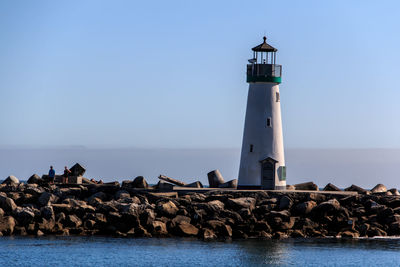 Lighthouse by sea against clear blue sky