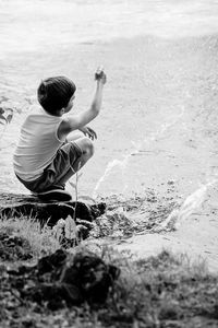 High angle rear view of boy playing with squirt gun while sitting on rock at lakeshore