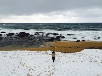 Boy walking at snow covered beach against sky