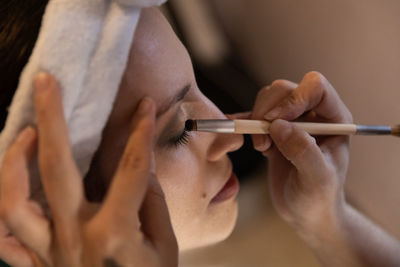 Cropped unrecognizable skilled beautician applying eyeshadow on female customer with eyes closed while working in beauty salon