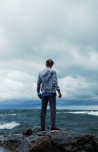 Rear view of man standing on rocky shore against cloudy sky