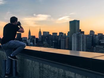 Rear view of man photographing city through camera against sky during sunset