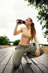 Portrait of young woman drinking water while standing against trees