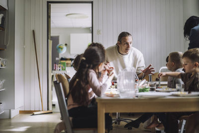 Male teacher talking with students having breakfast at table in day care center