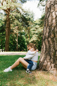 Young caucasian mother hugs her three year old son while sitting in the park near a tree 