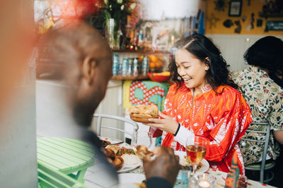 Smiling young woman eating lunch with friend sitting at table in restaurant seen through window