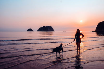 Silhouette mid adult woman with dog standing at beach against sky during sunset