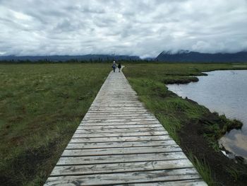 Footpath leading to pier on grassy field against cloudy sky