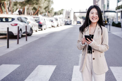 Smiling business woman listening to music on headphones and mobile phone crossing street in city