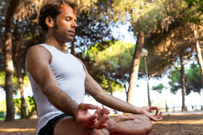 From below side view of barefoot focused male sitting in lotus pose with eyes closed on magenta mat and gesturing mudra during meditation session on sunny summer day in park