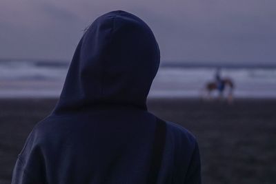 Rear view of person wearing hooded shirt at beach against sky during sunset