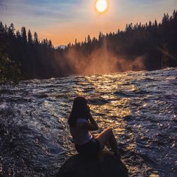 Rear view of woman sitting on rock at river during sunset