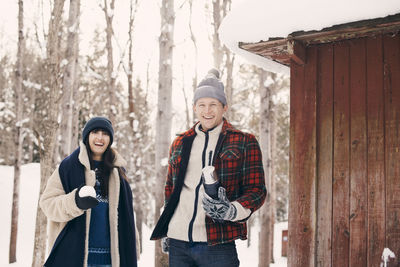 Portrait of happy friends holding snowball while standing by log cabin during winter