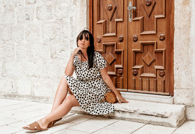 Beautiful young woman sitting in front of wooden door. summer, polka dot dress, lifestyle.