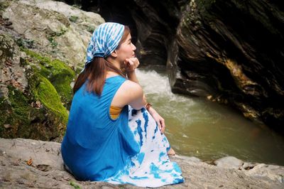 Thoughtful young woman looking away while sitting on rock by river in forest