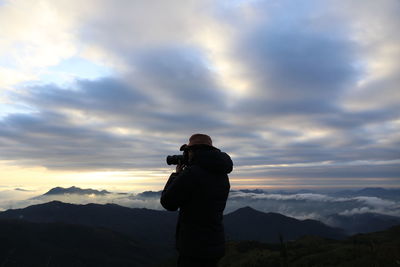 Person photographing by mountain against cloudy sky during sunrise