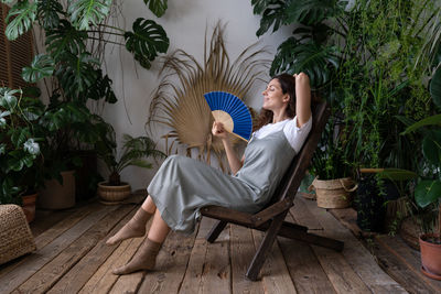 Young relaxed dreamy woman using paper fan while relaxing in beautiful refreshing home garden