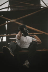 Low angle view of man holding camera while sitting against railing