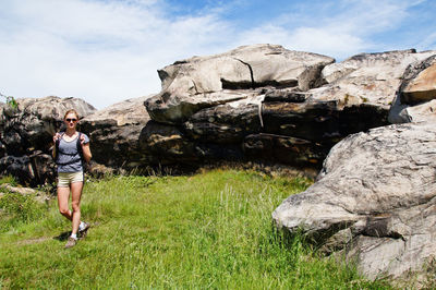 Female hiker walking on grassy field by rock formation against sky during sunny day