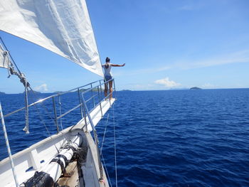 Rear view of young woman with arms outstretched sailing on sea against blue sky