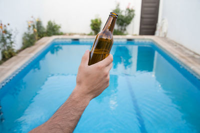 Cropped hand holding bottle against swimming pool