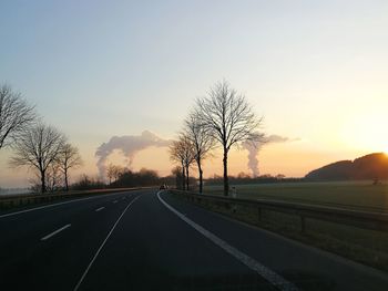 Road by bare trees against sky during sunset