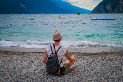 Rear view of woman looking at lake by mountains while sitting on pebbles