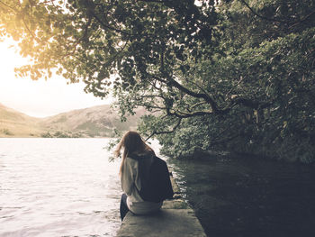 Woman sitting at lake against trees and sky