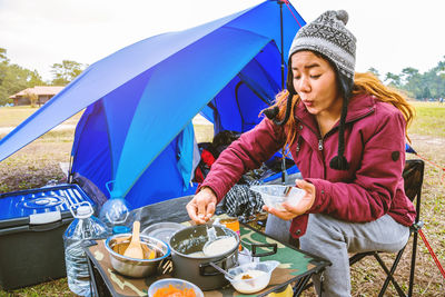 Woman wearing knit hat eating food sitting by tent outdoors