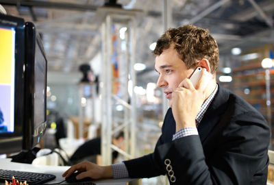 Side view of businessman talking on phone while using computer at office