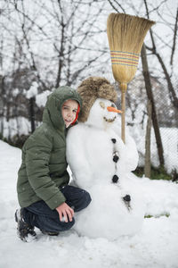 Full length of smiling boy crouching by snowman in yard during winter