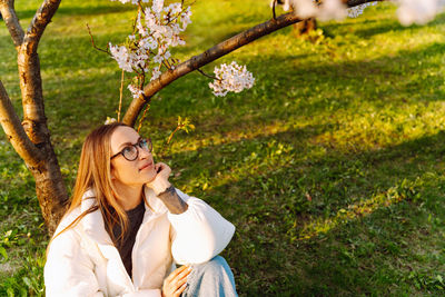 Beautiful young woman in white jacket and glasses sitting near blooming tree