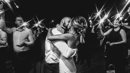 People looking at bride and bridegroom kissing during night