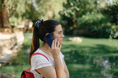 Medium shot of a young woman with a white dress and a red backpack talking on the phone in a park