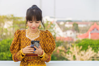 Asian woman wearing a yellow thai patterned dress watching her cell phone in her hands 