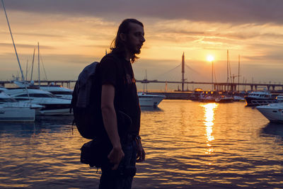 Guy in silhouette standing on beach with camera where yachts docked at the pier at sunset in summer