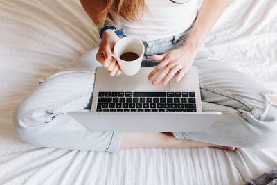 Low section of woman drinking coffee while using laptop on bed at home
